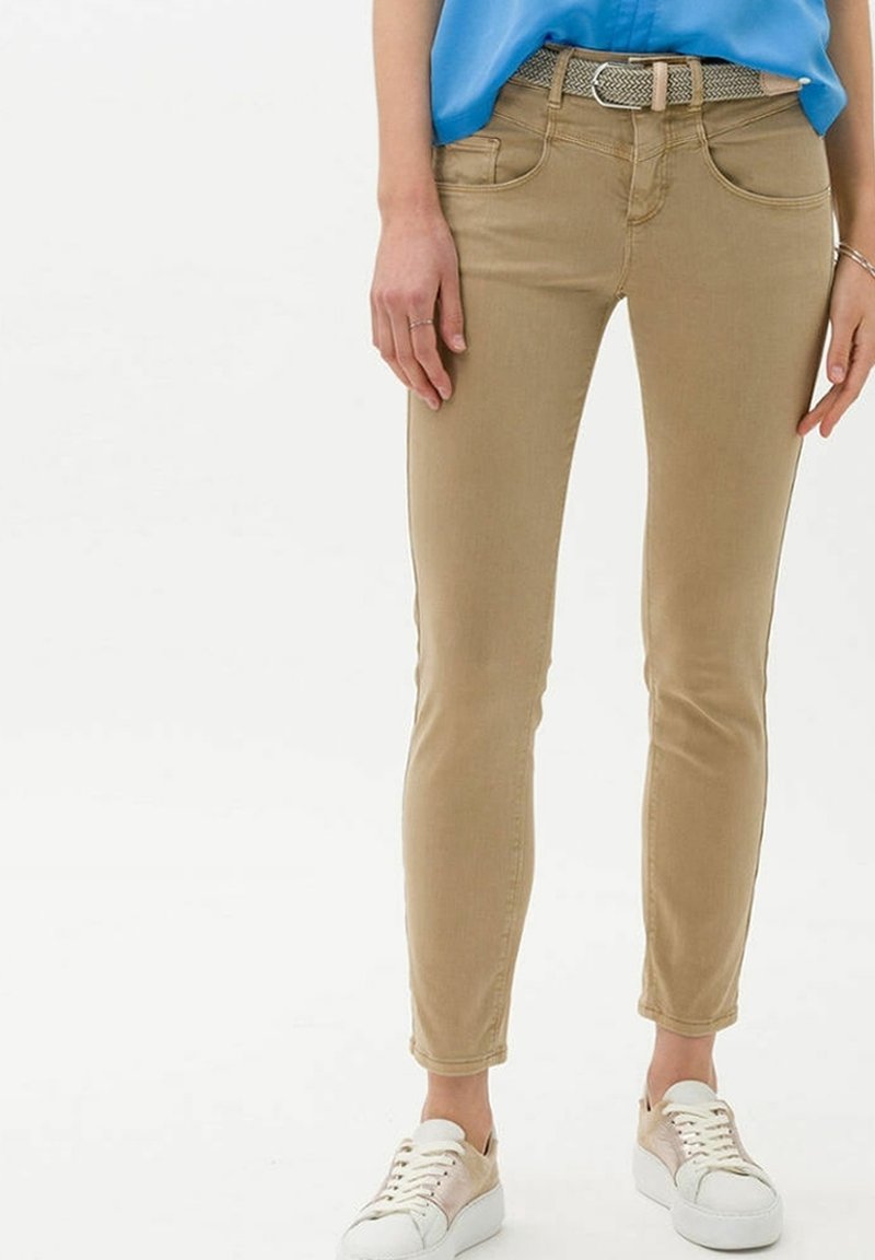 Femme portant un pantalon beige slim, ceinture tressée, chemise bleue et baskets blanches, debout, les bras détendus le long du corps.