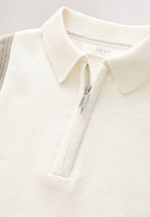 Flødefarvet pullover med halvlange lynlås, ribbet krave og blød tekstur. Har en kontrasterende stribe på skulderen.