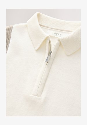 Kremine spalvos megztinis su užtrauktuku iki pusės, ribbinto audinio apykakle ir minkšta tekstūra. Pečių srityje yra kontrastinė juostelė.