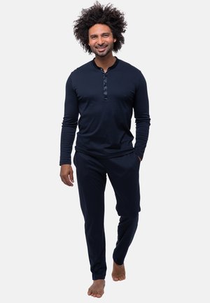 Mann mit lockigem Haar, der lächelt, barfuß geht und ein marineblaues Langarm-Henleyshirt sowie passende Hosen vor einem schlichten Hintergrund trägt.