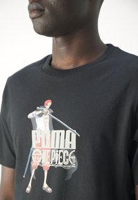 Camiseta de algodón negra que presenta un gráfico de un personaje con cabello rojo, sosteniendo una espada, con el texto "PUMA" y "ONE PIECE" en letras gruesas.