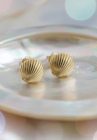 Boucles d'oreilles en forme de coquillage dorées avec des détails striés, posées sur une surface réfléchissante avec des reflets doux et irisés en arrière-plan.