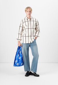 Camisa branca com padrão de quadrados castanhos, jeans azul claro, sapatos pretos e uma mochila azul com design floral branco. Modelo em pé contra um fundo simples.