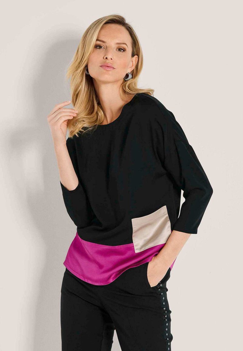 Frau mit blonden Haaren, die ein schwarzes Oberteil mit beigem Tascheneinsatz und pinkem Saum sowie schwarze Hosen mit seitlichen Knöpfen trägt, posiert vor einem hellen Hintergrund.