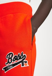 Orange sweatpants med ribbad midja, sidoficka och en svart broderad logotyp som står "BOSS" med sportiga accenter.
