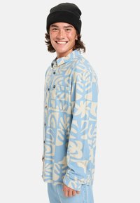 Blått flanellskjorta med krämfärgade abstrakta mönster, bröstficka och knapp-up-design. Bärs med en svart stickad mössa för att skapa en lager-på-lager-look.