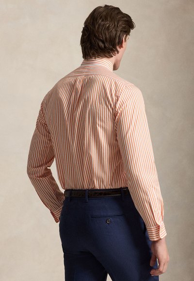 Homme de dos portant une chemise à manches longues rayée orange et blanche rentrée dans un pantalon bleu marine avec une ceinture noire.