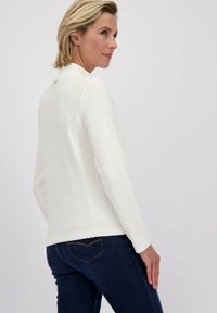 Femme avec les cheveux blonds courts, portant un haut à manches longues blanc texturé et un jean bleu foncé, de profil devant un fond uni.