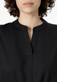 Black collared top featuring a front placket with a short slit. Made from a smooth fabric, it has a simple, clean design with visible stitching.