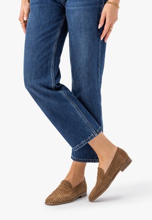 Osoba ubrana w niebieskie dżinsy i brązowe ażurowe mokasyny z zamszu, stojąca z nogami skrzyżowanymi na białym tle.