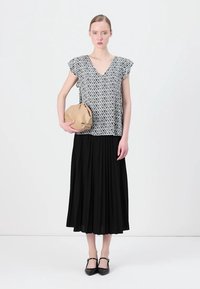 Svart plisserad kjol, mönstrad vit blus med korta ärmar och en beige kuvertväska. Modellen har på sig svarta skor; slätt tyg och lös passform.