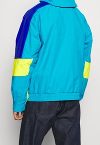 Kolorowa kurtka wiatrówka z turkusowymi, niebieskimi i żółtymi panelami, wykonana z lekkiego materiału, z elastycznymi mankietami i ściągaczem u dołu zapewniającymi dopasowanie.