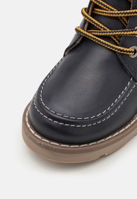 Laivaston siniset nahkakengät, joissa on kontrastiväriset valkoiset päällitikkausompeleet, pyöreä kärki, teksturoitu pinta ja keltaiset nauhat. Ruskea kumipohja.