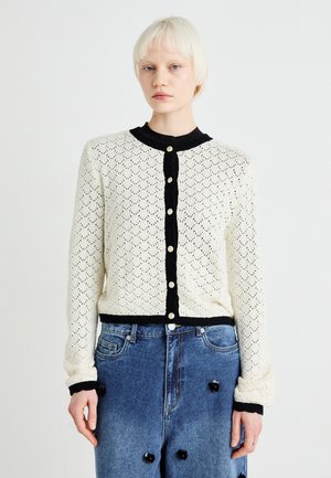 Cárdigan de punto crema con acentos negros de canalé, cierre de botones y un patrón similar al encaje. Combinado con jeans de mezclilla azul adornados con adornos.