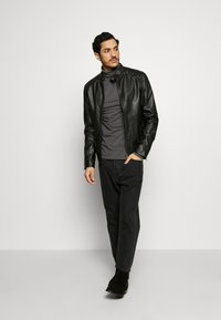 Schwarze Lederjacke mit Mandarin-Kragen, tailliertem Schnitt und Schulterdetails. Getragen über einem grauen Rollkragenpullover und kombiniert mit dunklen Hosen.