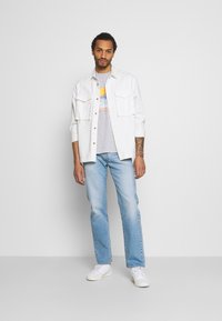 Camisa blanca de botones sobre una camiseta gráfica gris, combinada con jeans de un azul claro y zapatillas blancas. Conjunto simple y casual con bolsillos.