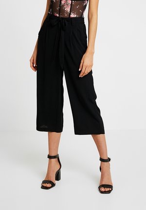 Mujer con pantalones negros de pierna ancha de talle alto y corte cropped con cinturón anudado, combinados con sandalias negras de tacón cuadrado con tira al tobillo.