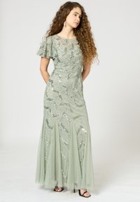 Langes, hellgrünes Kleid mit pailettenbesetztem Blättermuster, durchsichtigen, ausladenden Ärmeln und einem fließenden Rock, kombiniert mit schwarzen Riemchenschuhen.