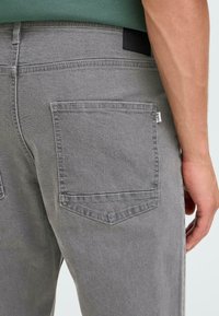 Sivi jeans s teksturo, ki je gladka, z dvema žepoma na zadnji strani in črno oznako na pasu. Prisotne so preproste šive.