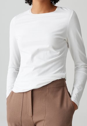 Femme portant une chemise blanche ajustée à manches longues rentrée dans un pantalon marron taille haute, les mains dans les poches.