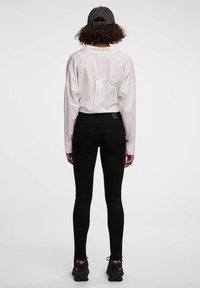 Chemise blanche à manches longues, jean skinny noir et baskets noires. Vue de dos, montrant le col de la chemise et les poches du jean. Texture lisse.