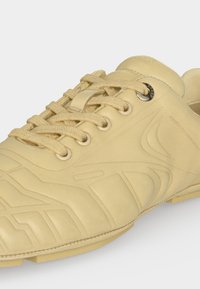 Baskets beiges avec motifs texturés, fermeture à lacets avec lacets beiges assortis, œillet métallique et design bas sur un fond neutre.