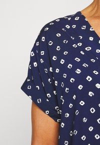Blusa azul-marinho com decote em V e mangas curtas, adornada com padrões quadrados brancos, mostrando um tecido suave e texturizado.
