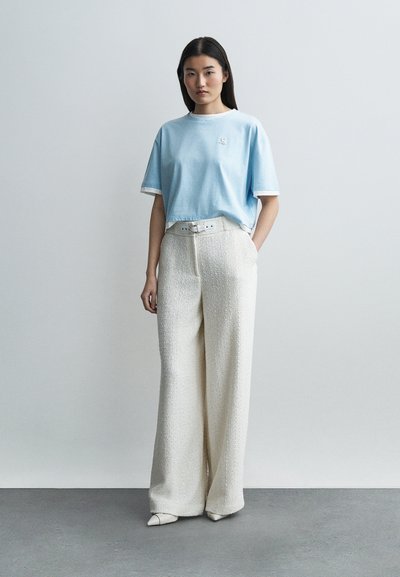 Γυναίκα στέκεται μπροστά σε απλό φόντο φορώντας ανοιχτό μπλε φαρδιά μπλούζα και ψηλόμεσα λευκά παντελόνια με φαρδιά μπατζάκια, ζώνη και λευκά παπούτσια.