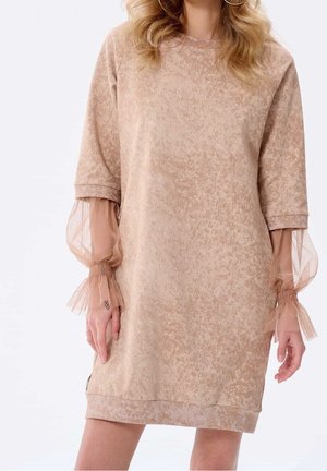 Beige tekstureret kjole med gennemsigtige puffede lange ærmer samlet ved håndleddene, båret af en person med blondt, bølget hår og store hoop-øreringe.