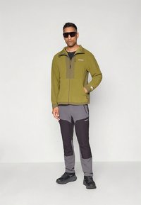 Chaqueta de forro polar verde oliva con cierre de cremallera y panel contrastante en color beige; pantalones negros y grises con diseño en bloques de color; zapatos negros; gafas de sol.