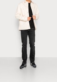 Ljus beige knappad skjorta över en svart t-shirt, tillsammans med mörka jeans och svarta Nike-sneakers med vita detaljer.