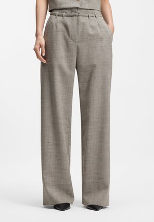 Nainen, joka pukeutunut korkeavyötäröisiin, leveälahkeisiin beigeihin housuihin, joissa on vyölenkit ja taskut, yhdistettynä kärjellisiin mustiin kenkiin, seisoo valkoista taustaa vasten.