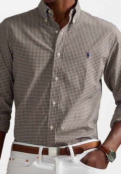 Brown and white checked shirt with button-down collar, featuring a blue logo embroidery. Paired with a brown belt and white pants.