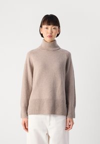 Donna che indossa un maglione beige a collo alto abbinato a pantaloni bianchi, in piedi davanti a uno sfondo grigio chiaro uniforme.