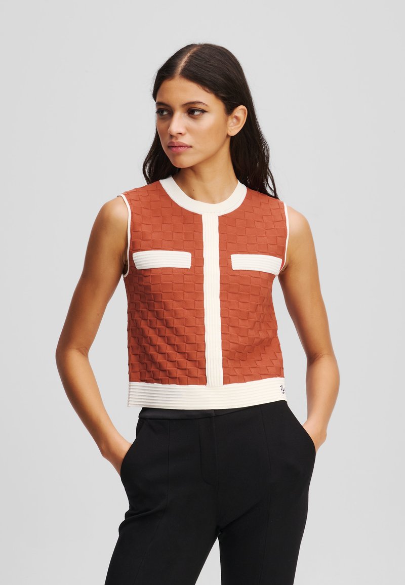 Ermeløs crop top i rust med et teksturert mønster, hvit ribbestrikket halslinning og horisontale striper i kanten; har to brystlommer.