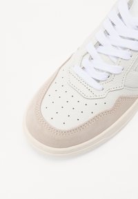 Sneaker bianca in pelle con accenti in suede beige chiaro, punta arrotondata, tomaia perforata e lacci bianchi su una suola chiara.