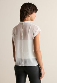 Hvid, halvgennemsigtig bluse med korte ærmer, høj halsudskæring og en nøglehul-detail bagtil, parret med sorte slim-fit bukser.