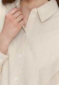 Camisa de botones con un patrón a rayas en beige y blanco. Presenta un cuello puntiagudo, botones blancos y mangas largas. Textura suave.