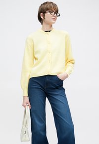 Κίτρινη πλεκτή ζακέτα με στρογγυλή λαιμόκοψη, κουμπιά μπροστά και ριμπ μανσέτες, συνδυασμένη με σκούρο μπλε τζιν παντελόνι με φαρδιά μπατζάκια και λευκή τσάντα.