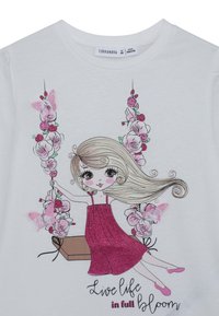 T-shirt bianca con una grafica di una ragazza in un vestito rosa glitterato su un'altalena, con accenti floreali e farfalle. Il testo recita "Vivi la vita in piena fioritura."