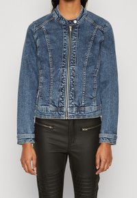 En blå denimjacka med hög krage, dragkedjestängning och vertikala sömdetaljer. Buren över svarta läderbyxor med texturerade accenter.