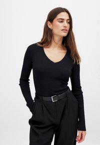Black ribbed long-sleeve top with a V-neck design, textured fabric, and fitted silhouette, paired with high-waisted black pants and a belt.