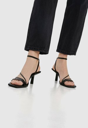 Sandales noires à talons avec des fines lanières entrecroisées et un bout carré. Elles présentent un talon mince et effilé ainsi qu'une texture lisse et brillante.