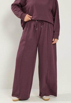 Persoon draagt losse, wijdvallende bordeauxrode broek met elastisch koord in de taille en bijpassend longsleeve top, staand met één hand in de zak.