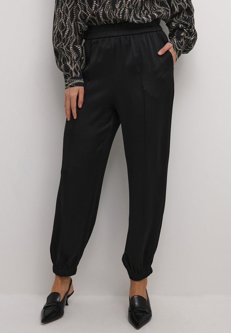 Pantaloni jogger in satin nero con vita elastica, caviglie a polsino e una texture liscia, indossati con mule a punta e una blouse con motivo.