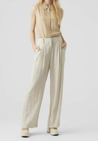 Beżowa bluzka bez rękawów z guzikami, zestawiona z jasnobeżowymi szerokimi spodniami. Modelka ma na sobie kremowe buty na platformie, ręce w kieszeniach.