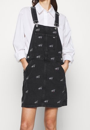 Frau trägt ein schwarzes Denim-Latzkleid mit weißen Signatur-Logos und Vordertasche über einem weißen, langärmligen, geknöpften Hemd, Hände in den Taschen.