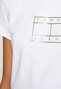 Bílý bavlněný tričko s zlatým obdélníkovým logem, uvnitř kterého jsou slova "TOMMY" a "JEANS". Krátké rukávy a volný střih.