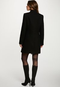 Abrigo negro con una silueta estructurada, cuello alto y mangas largas. Combinado con medias transparentes y botas negras de textura hasta la rodilla.