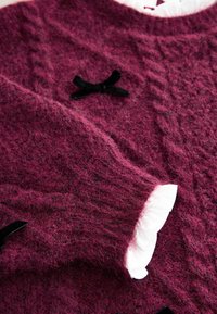 Weinroter Wollpullover mit strukturierter Oberfläche, schwarzen Samtschleifen als Akzente und weißen, gerafften Manschetten.